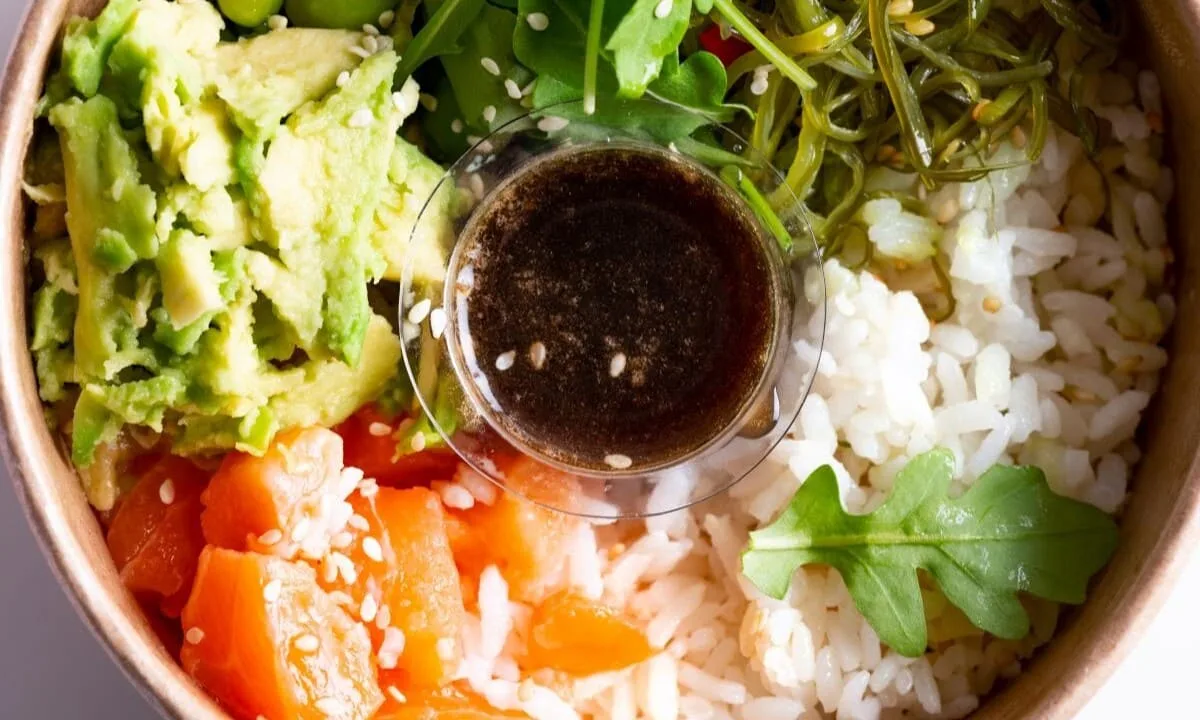Poke bowl with salmon, avocado, rice, seaweed, and a cup of soy-based poke sauce with sesame seeds.