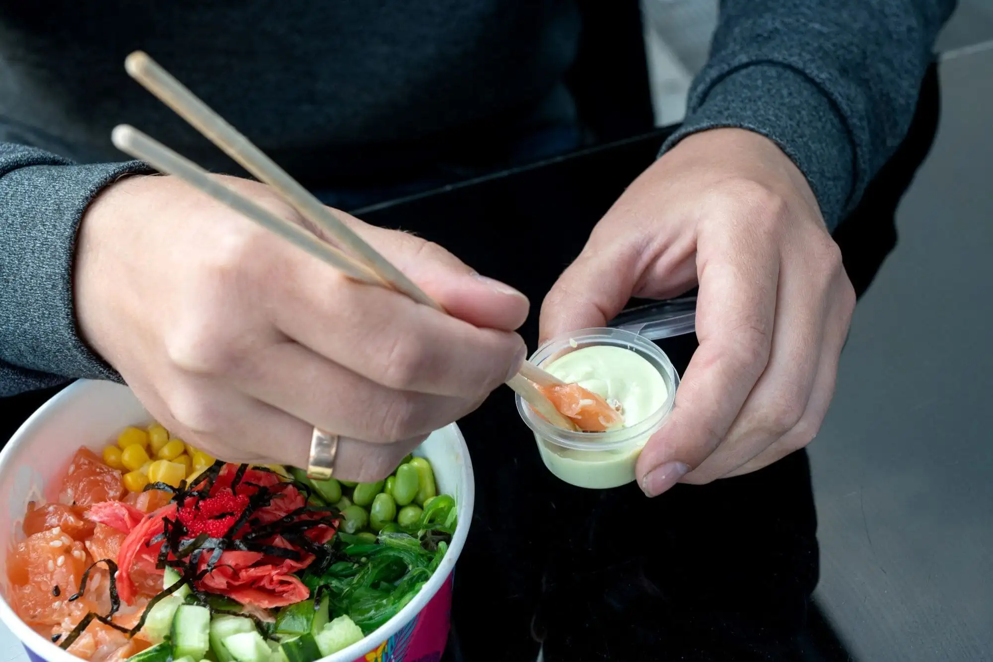 Person dipping chopstick in sauce, preparing colorful poke bowl with salmon, corn, edamame, and seaweed.