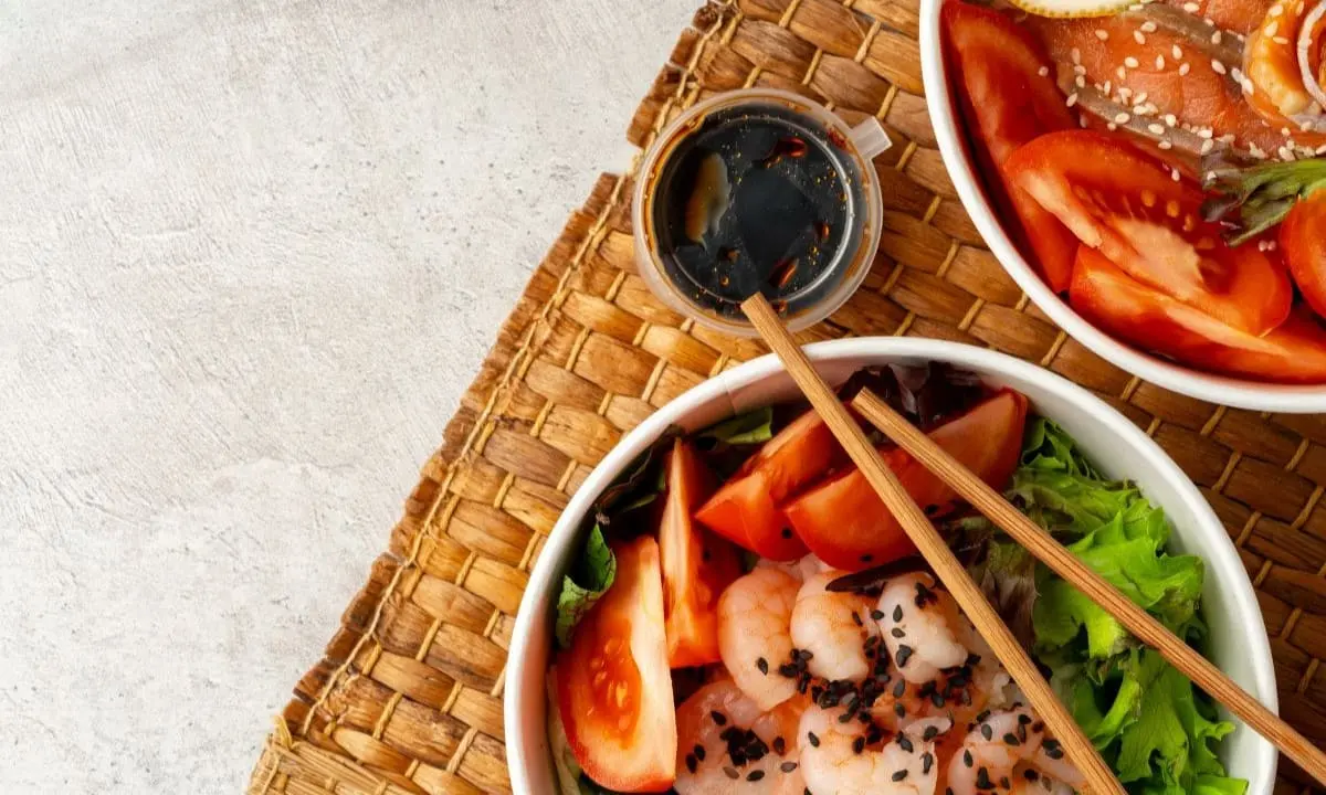 Traditional poke bowl with shrimp, tomatoes, and sesame seeds, paired with soy-based poke sauce on woven mat.