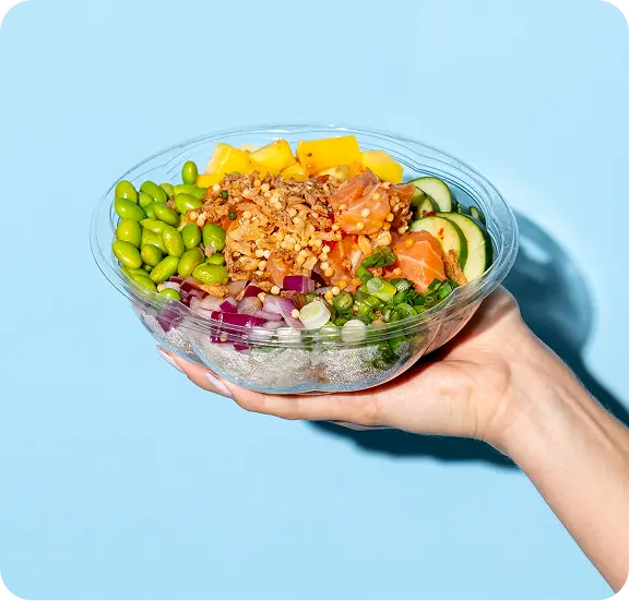 Hand holding clear bowl of poke-style salad with edamame, mango, salmon