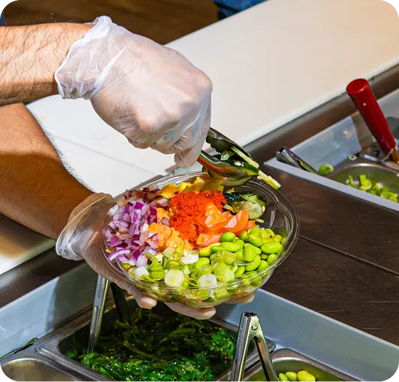 Gloved hands assembling a colorful poke bowl with edamame and orange fish roe