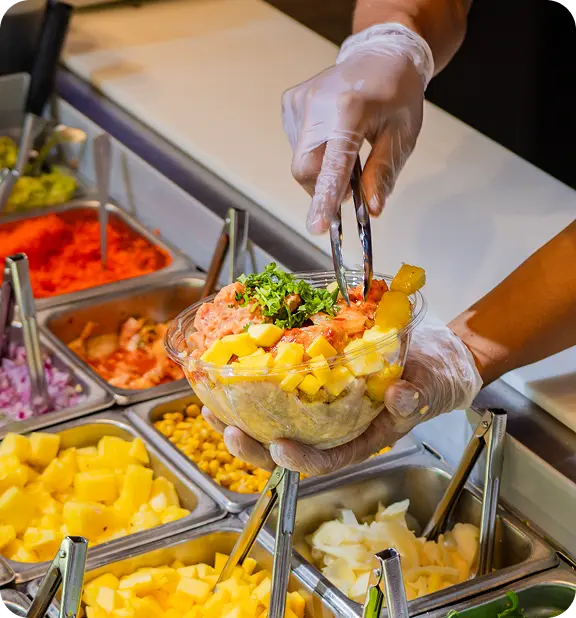 Gloved hands assembling poke bowl with diced salmon, mango, corn, cilantro